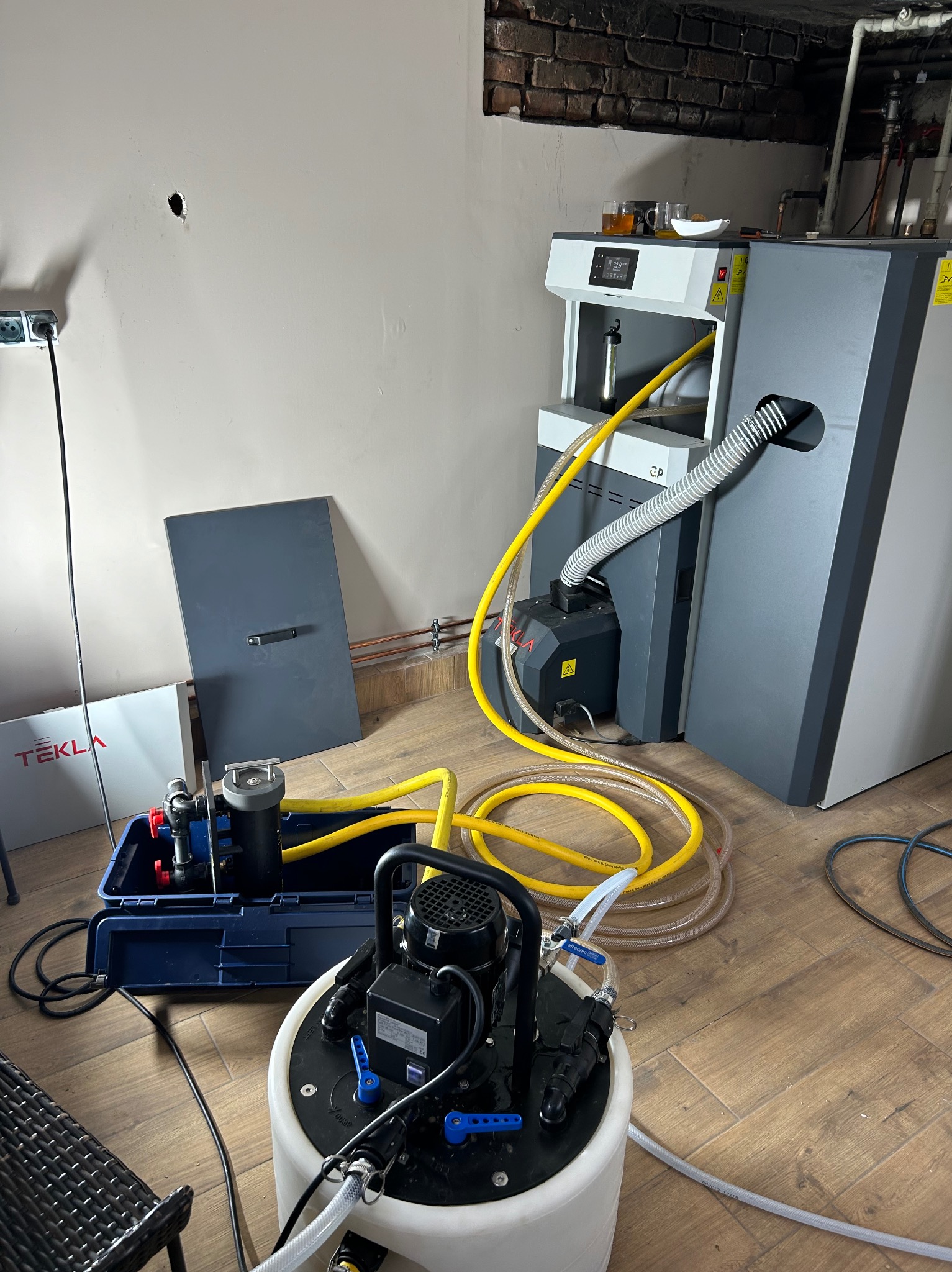 Nowoczesny piec CO z podajnikiem, obok urządzenia do testowania szczelności instalacji, widoczne rurki miedziane i otwór w ścianie.