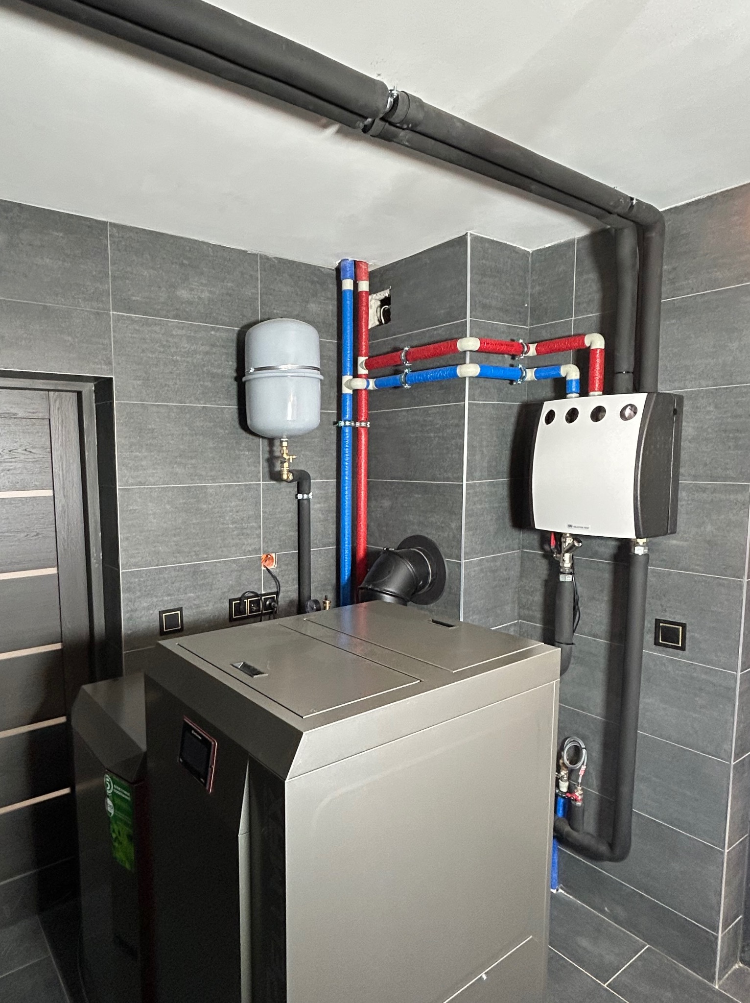 Nowoczesna kotłownia z szarą płytką ścienną, widoczny piec CO, zbiornik wyrównawczy, oraz instalacja z rur w kolorach niebieskim i czerwonym, czarne rury izolowane biegnące pod sufitem.