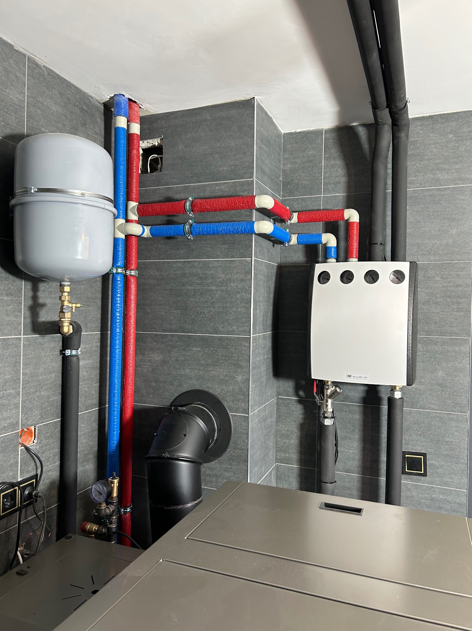 Kotłownia z szarymi płytkami: zbiornik wyrównawczy, rury grzewcze w izolacji czerwonej i niebieskiej, czarny komin i biały panel sterowania z manometrami.