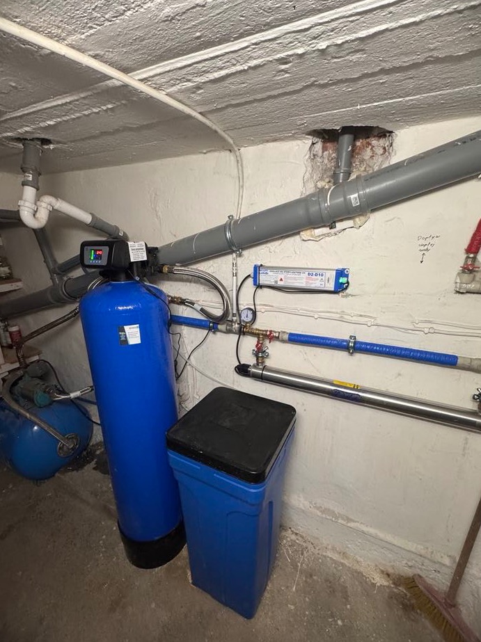 Niebieski filtr wody z czarnym panelem sterowania i zbiornik na sól w piwnicy, widoczne rury instalacji wodnej i fragment instalacji elektrycznej.