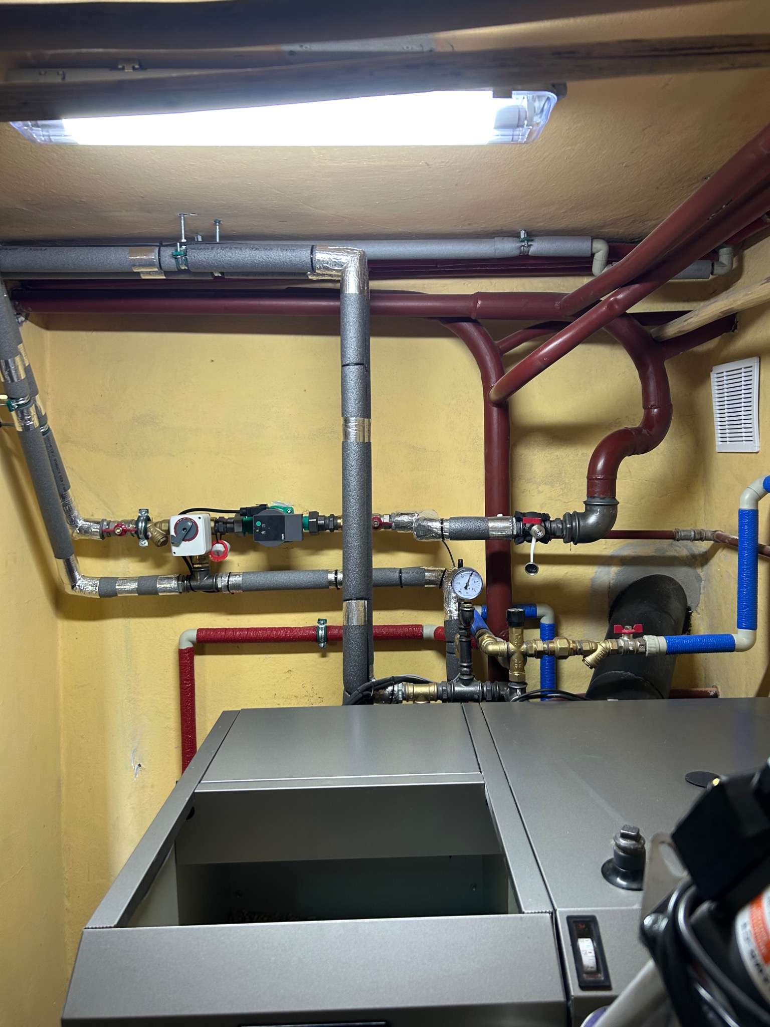 Złożona instalacja CO z rur miedzianych i zaworów, z manometrem i pompą obiegową. Szare rury izolowane termicznie, żółte ściany. Widoczny piec CO.