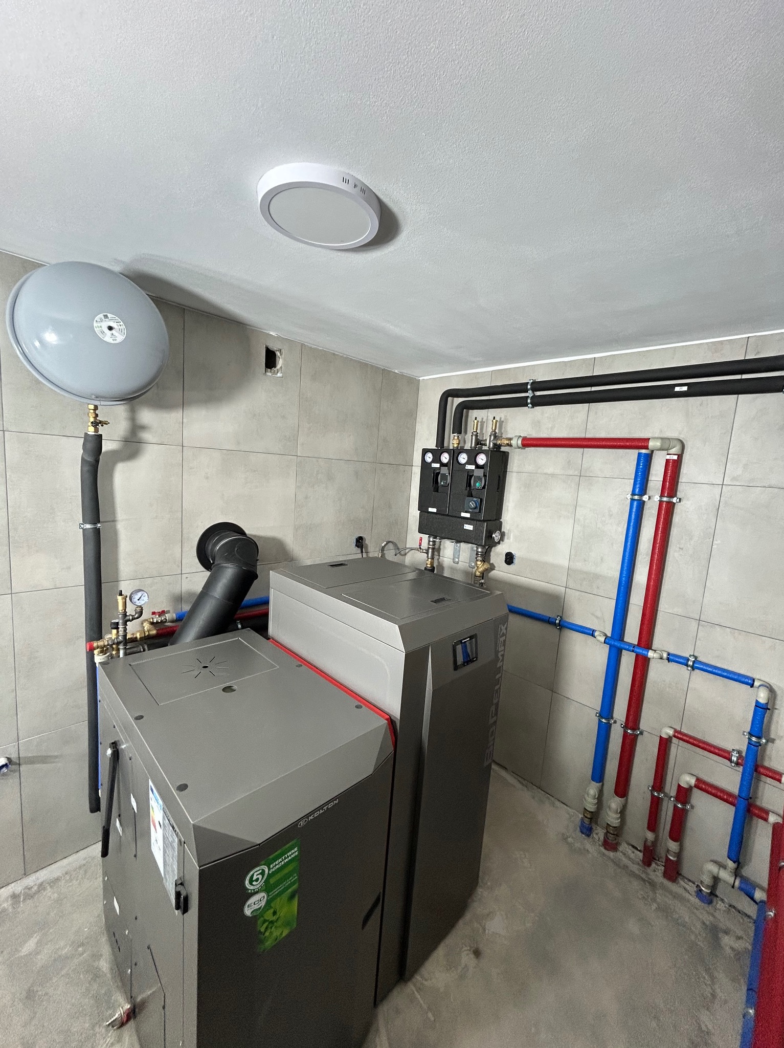 Nowoczesny piec na pellet w kotłowni, z widocznymi rurami instalacji grzewczej w kolorach czerwonym i niebieskim, na tle szarych płytek ściennych.