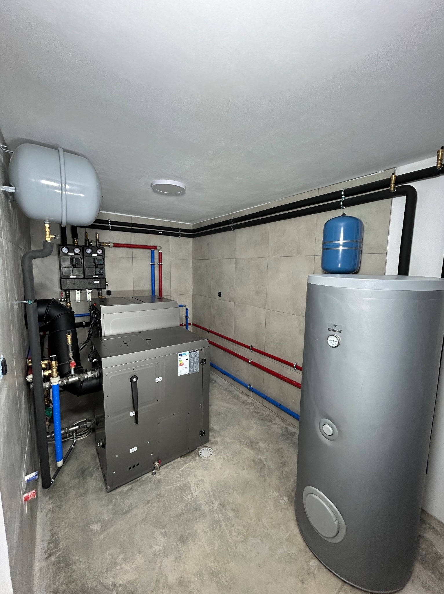 Nowoczesna kotłownia z szarym bojlerem i pompą ciepła. Widoczne rury w kolorach niebieskim i czerwonym. Ściany wyłożone płytkami w odcieniach szarości.