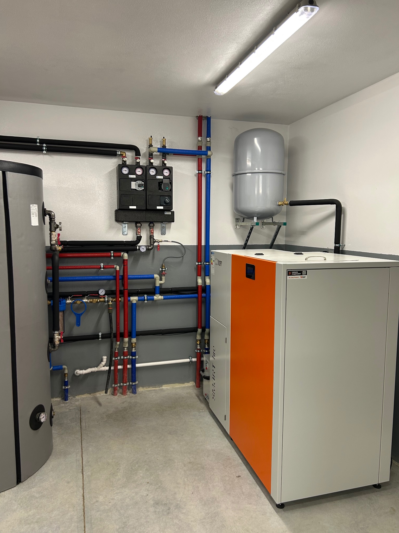 Zmontowana kotłownia z piecem CO z pomarańczowym akcentem, zbiornikiem wyrównawczym i kompletną instalacją grzewczą z rurami w kolorach czerwonym i niebieskim.