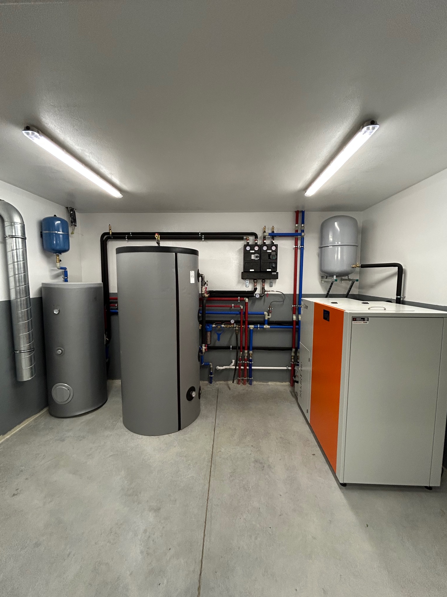 Kotłownia z instalacją grzewczą i zbiornikami w Ostrowsku. Widoczne rury, zawory i osprzęt. Nowoczesny piec CO z pomarańczowym akcentem. Szare ściany i betonowa posadzka.