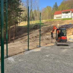 Wkopane słupki ogrodzeniowe w ziemię, widoczny mały traktor z wiertłem, przygotowanie do montażu ogrodzenia na nierównym terenie, dom w tle.