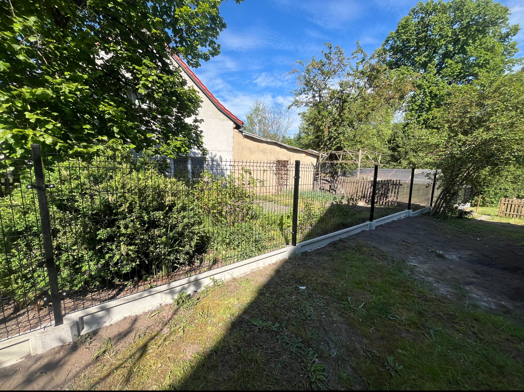 Czarny panelowy system ogrodzeniowy na betonowej podmurówce, widok z zewnątrz posesji, zarośnięte krzewy przy ogrodzeniu, w tle budynek gospodarczy z czerwonym dachem.