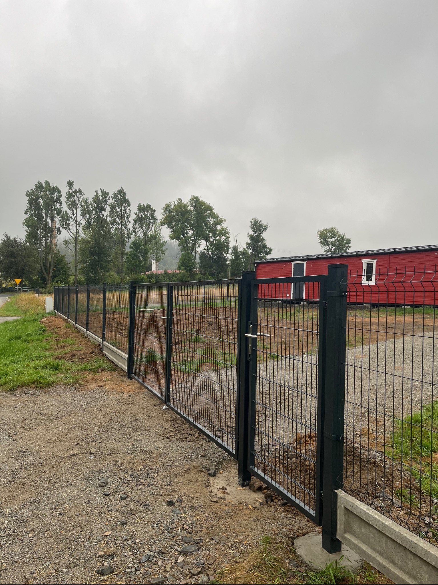 Nowoczesne, czarne ogrodzenie panelowe z bramką, osadzone na betonowej podmurówce. W tle czerwony budynek i drzewa w mglisty dzień.