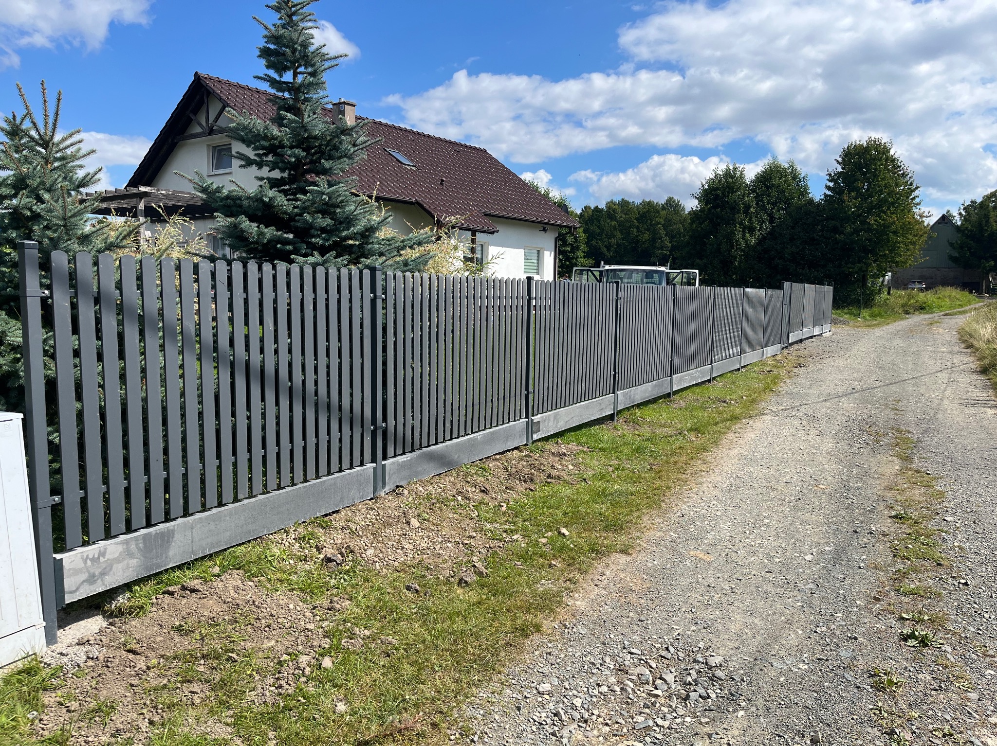 Nowoczesne ogrodzenie palisadowe w kolorze grafitowym, z betonową podmurówką, na tle domu jednorodzinnego i błękitnego nieba. Widok z perspektywy ścieżki.