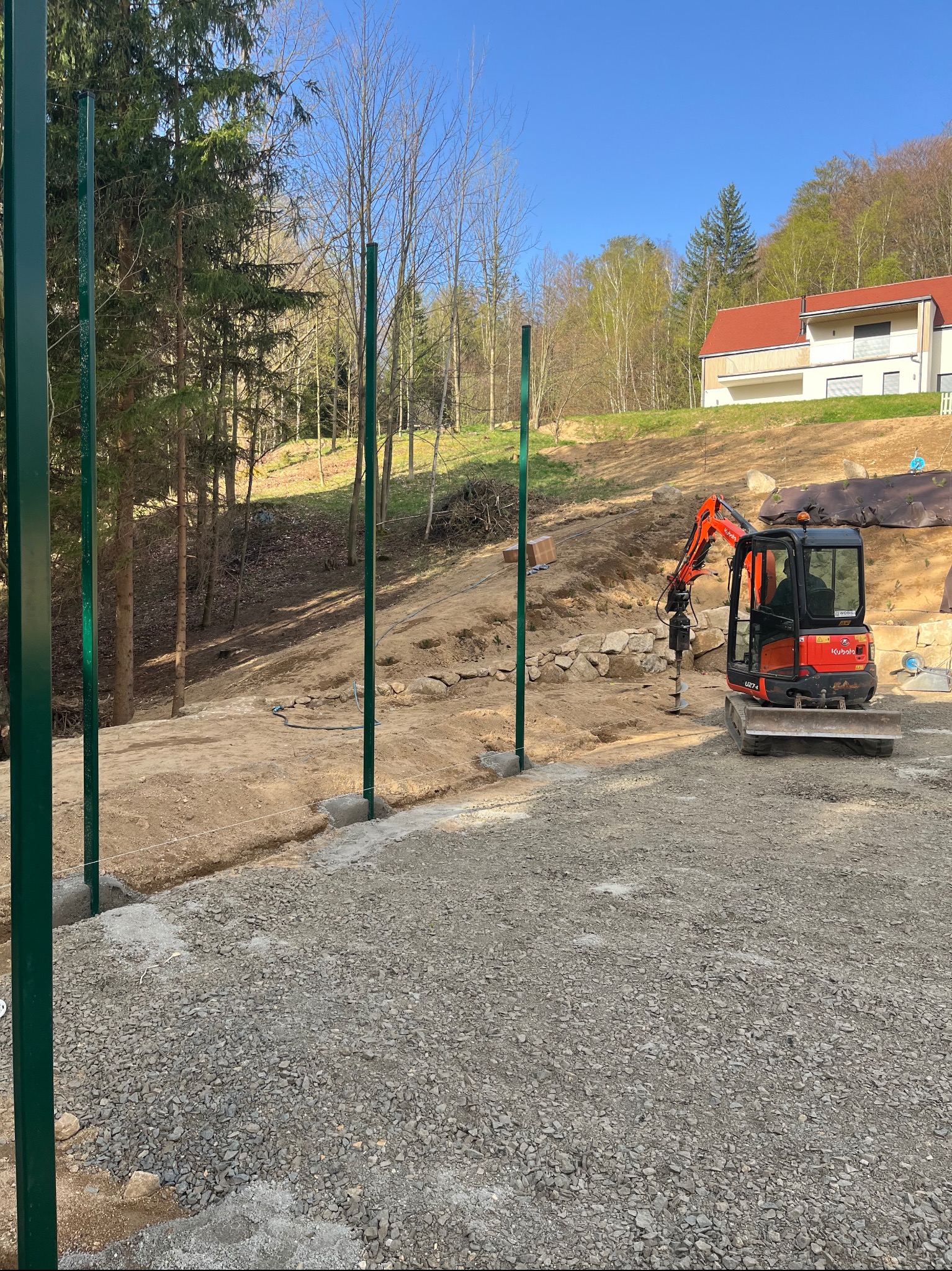 Wkopane słupki ogrodzeniowe w ziemię, widoczny mały traktor z wiertłem, przygotowanie do montażu ogrodzenia na nierównym terenie, dom w tle.
