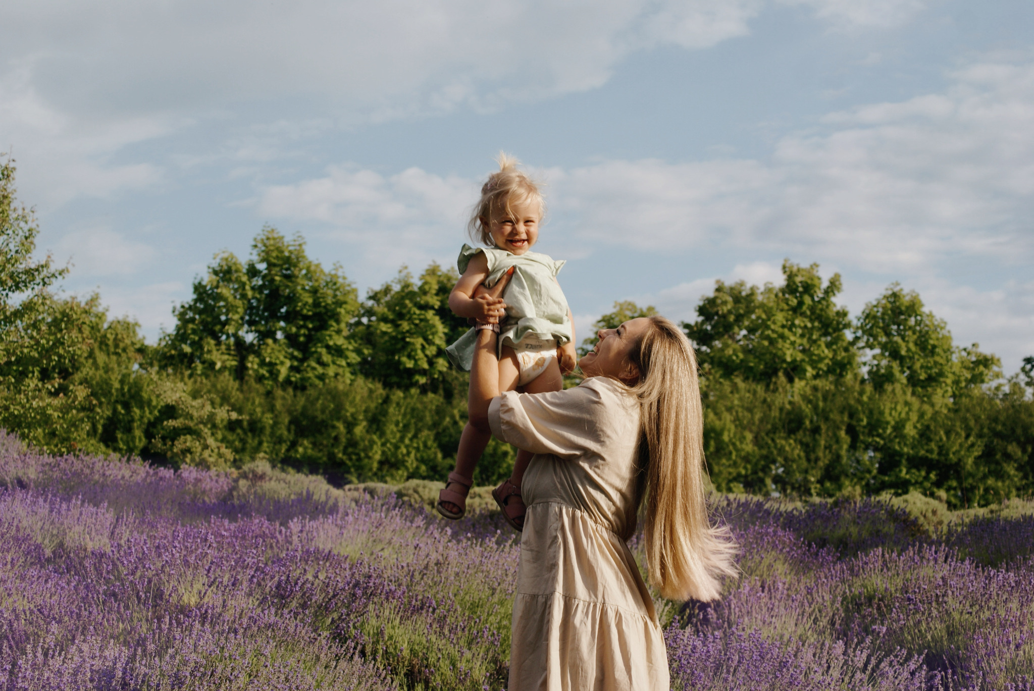 Uśmiechnięta matka podnosi w górę roześmianą córeczkę na tle kwitnącego pola lawendy i zieleni drzew.