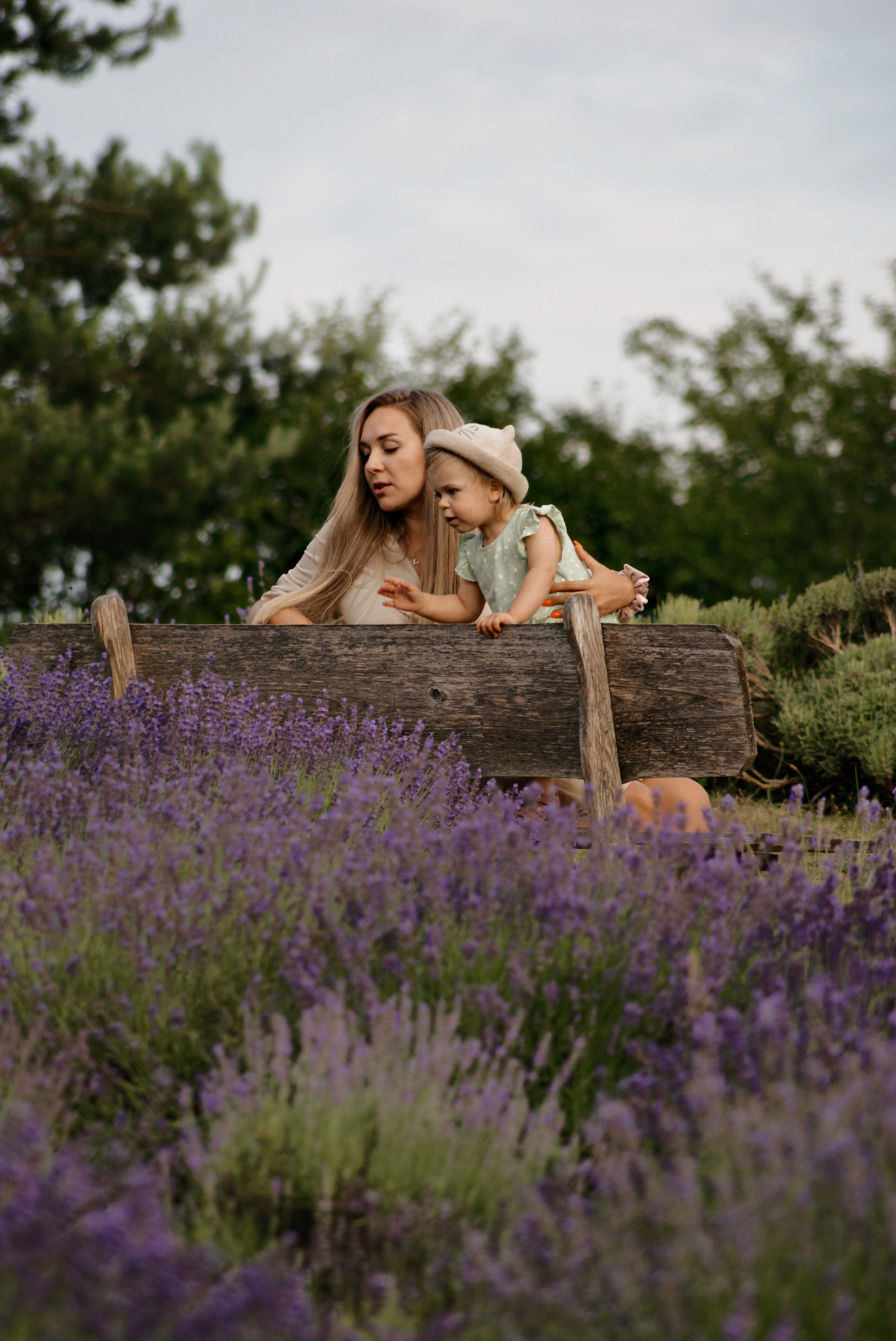 Matka z małą córeczką w kapeluszu siedzą na drewnianej ławce otoczonej polem kwitnącej lawendy, z zalesionym tłem.