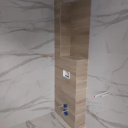 Nowoczesna łazienka w trakcie wykańczania: marmurowe płytki ścienne, zabudowa z półkami wyłożona płytkami imitującymi drewno, widoczne rury instalacji sanitarnej i podtynkowy system spłukiwania.