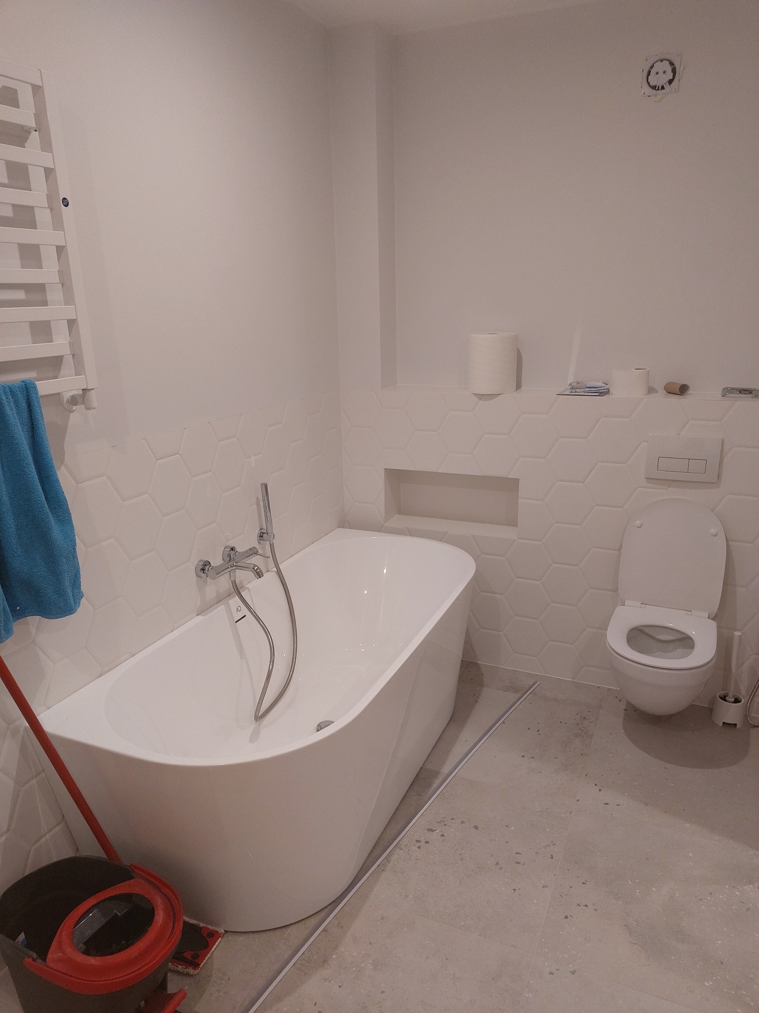 Nowoczesna łazienka z wolnostojącą wanną, heksagonalnymi płytkami na ścianie, podwieszaną toaletą i grzejnikiem drabinkowym z niebieskim ręcznikiem, widoczne ślady po remoncie: wiadro z mopem.