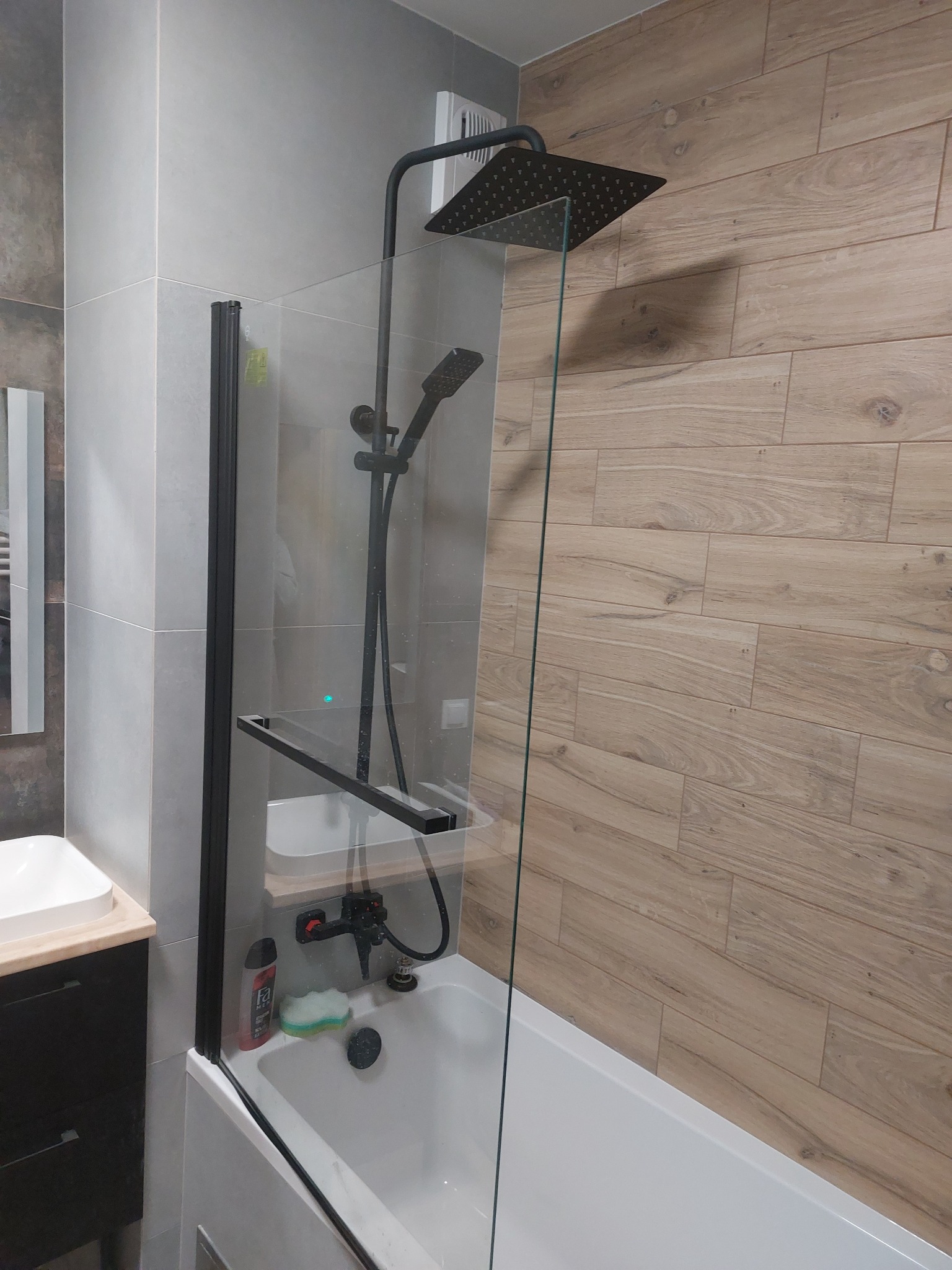 Nowoczesna łazienka z kabiną prysznicową typu walk-in z czarnymi profilami, deszczownicą i baterią w kolorze czarnym, jasne drewnopodobne płytki na ścianie, umywalka nablatowa z szafką.