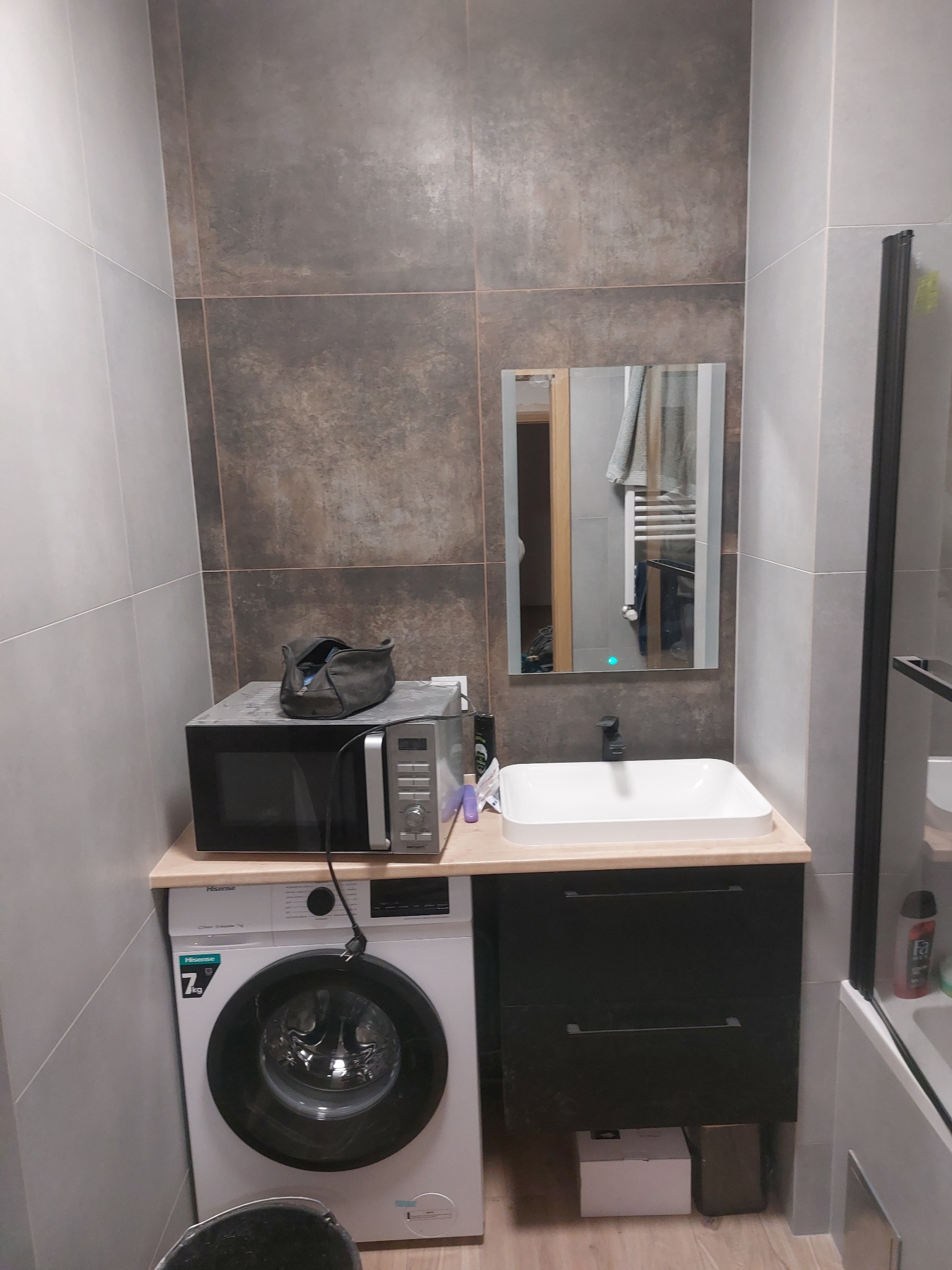 Funkcjonalna łazienka z pralką pod blatem, na którym stoi mikrofalówka, umywalka nablatowa z czarną szafką i lustro z podświetleniem LED.