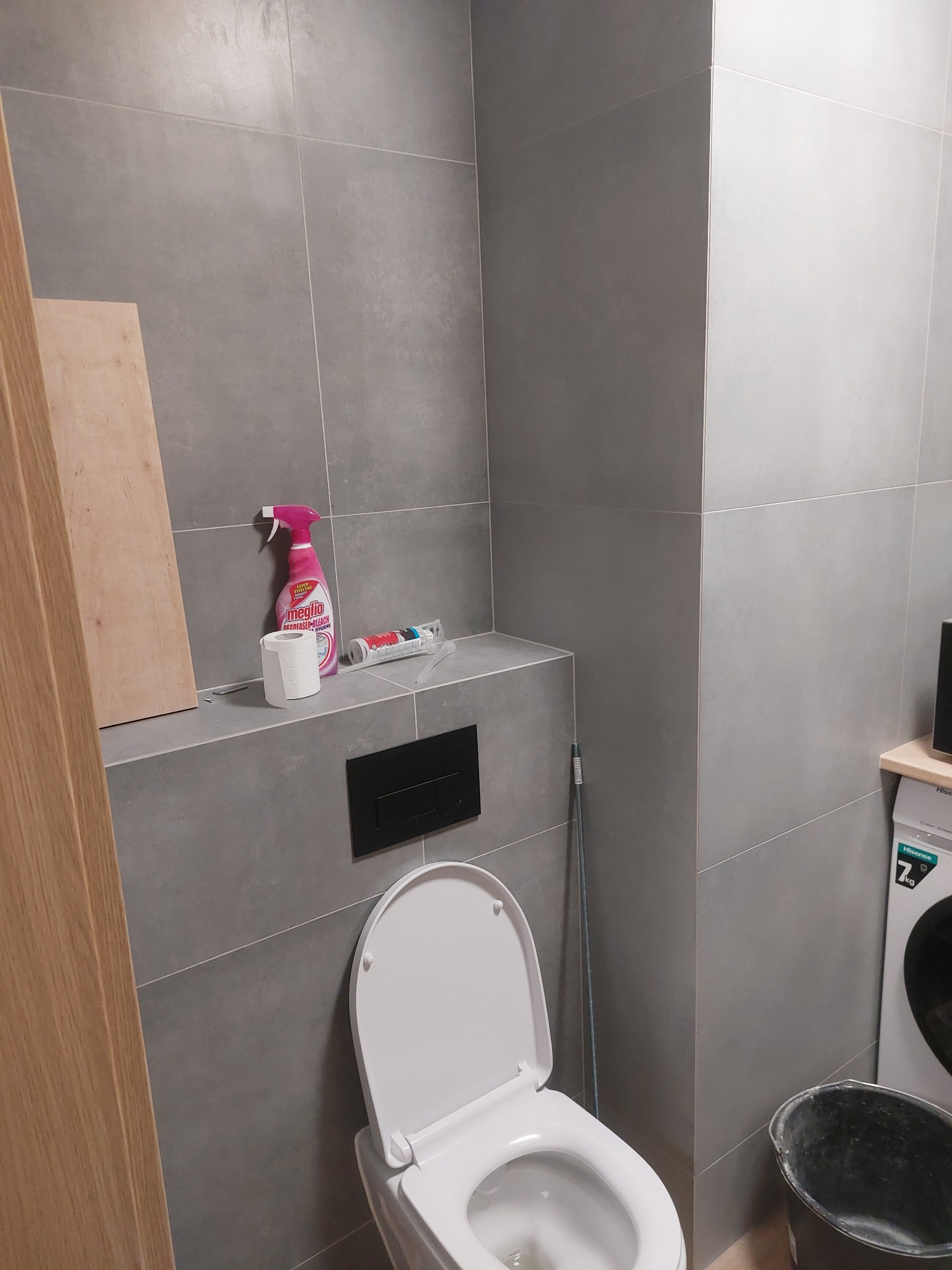 Szara łazienka w trakcie prac wykończeniowych, widoczna otwarta toaleta, pralka, wiadro, chemia do czyszczenia i deska.