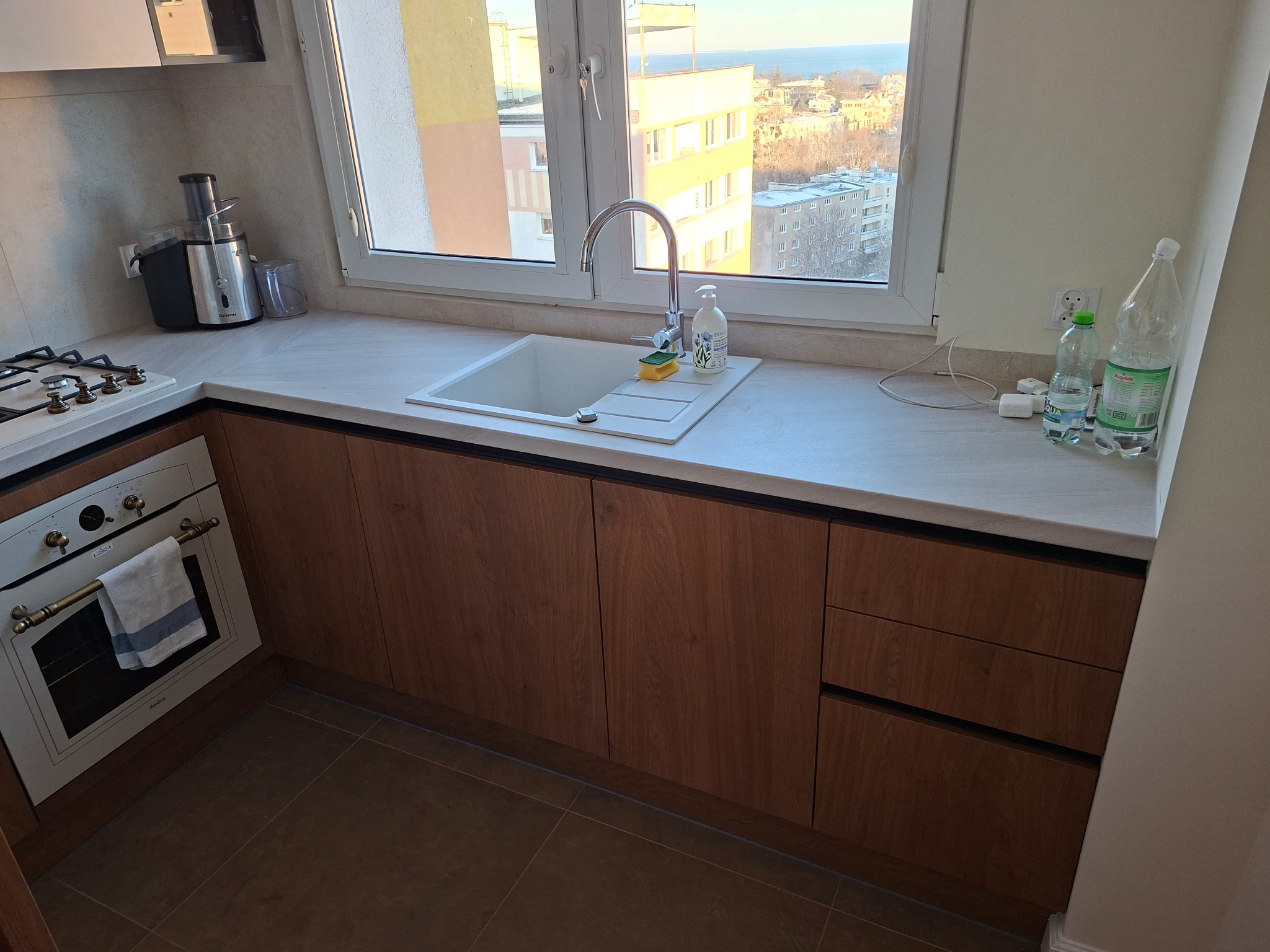 Nowoczesna kuchnia z drewnianymi szafkami, białym blatem i zlewozmywakiem pod oknem z widokiem na miasto i morze. Widoczny piekarnik i akcesoria kuchenne.