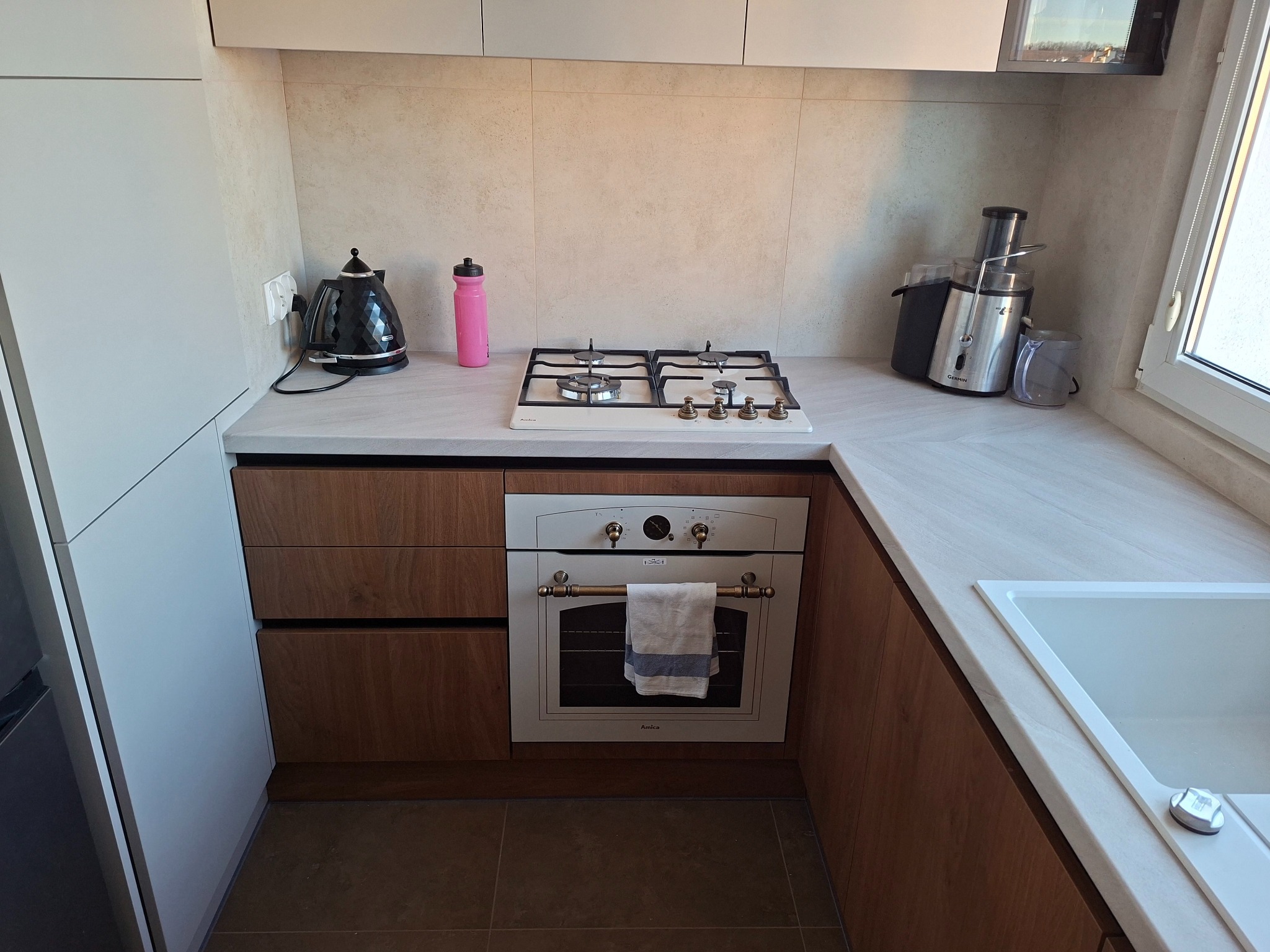 Nowoczesna kuchnia z drewnianymi szafkami, białym blatem i sprzętami AGD. Uwagę przyciąga piekarnik Amica z ręcznikiem na drzwiczkach i płyta gazowa.