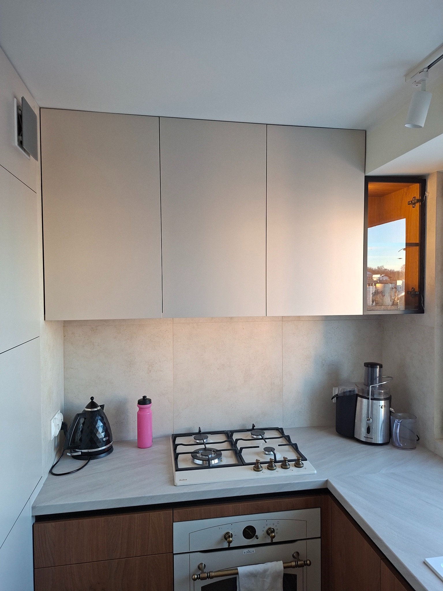 Jasna kuchnia z beżowymi szafkami, drewnianymi frontami dolnymi i blatem imitującym kamień. Widoczny sprzęt AGD i okno z widokiem na zewnątrz.