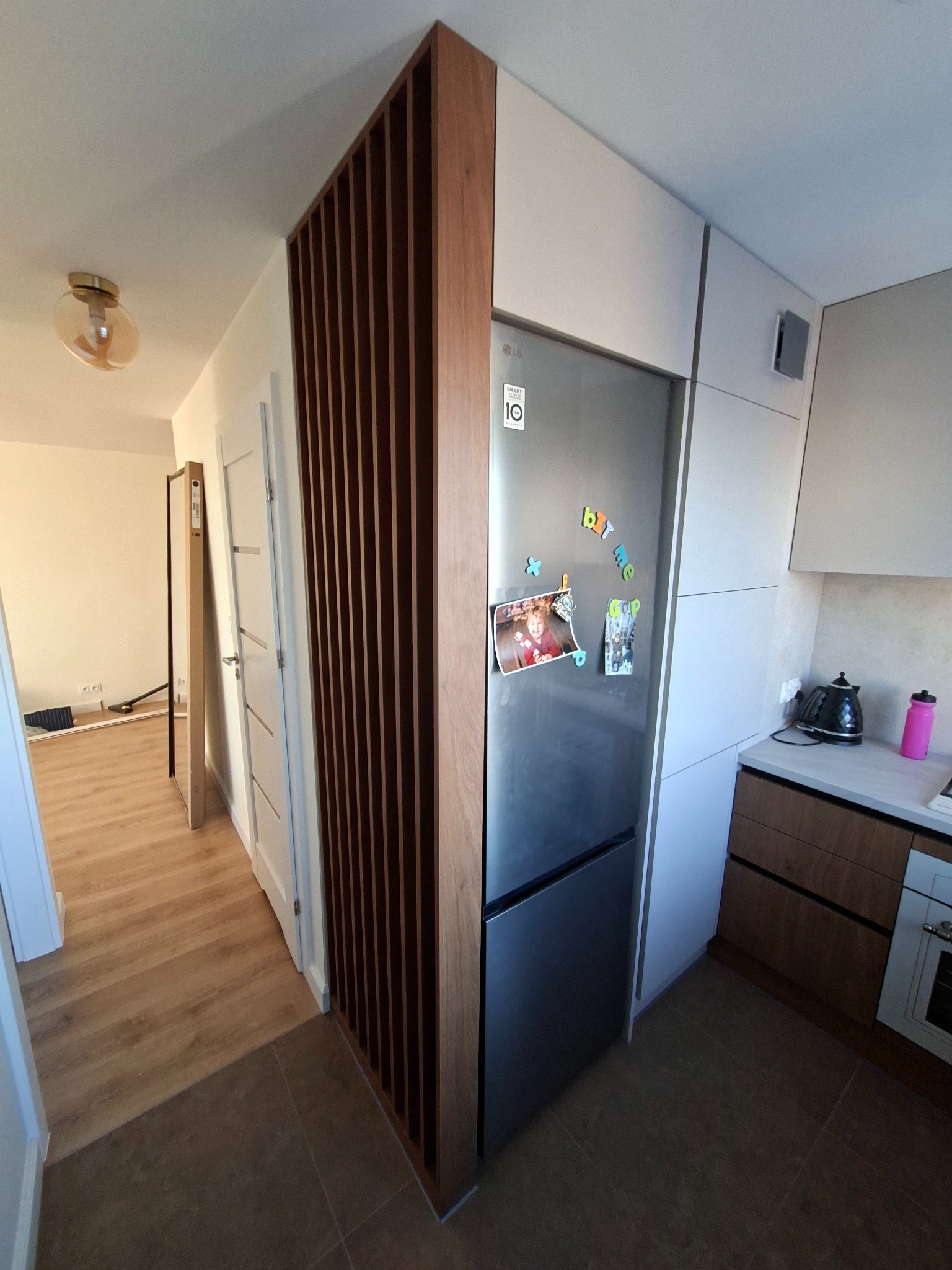 Nowoczesna kuchnia z drewnianą ścianką działową z pionowych listew, przylegająca do zabudowy lodówki. Widoczne białe fronty szafek i sprzęty AGD. Podłoga w odcieniach brązu.