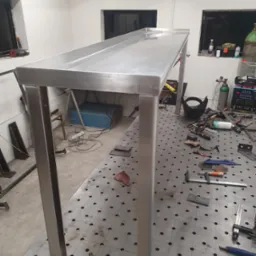 Stalowy stół warsztatowy z profili zamkniętych, spawany, na tle warsztatu z narzędziami i sprzętem spawalniczym.