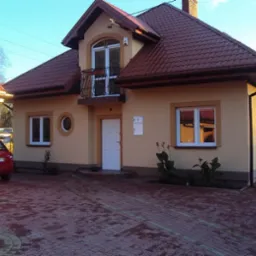 Jasna elewacja domu jednorodzinnego z czerwonym dachem, balkonem z kutą balustradą i obramowaniami okien w kolorze piaskowym, na pierwszym planie czerwona kostka brukowa.