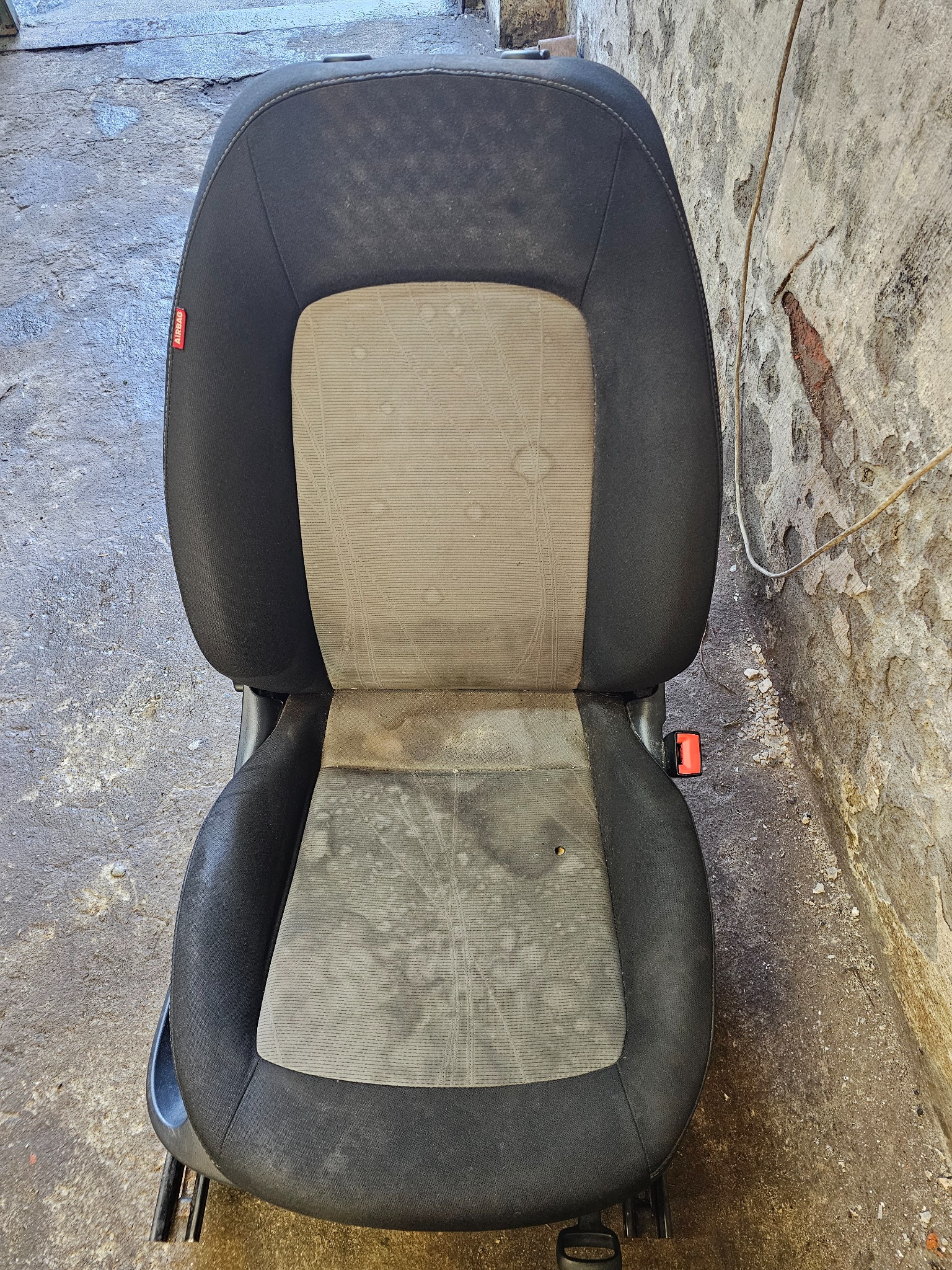 Brudny fotel samochodowy z widocznymi plamami na tapicerce, stojący na betonowej podłodze przy szorstkiej ścianie.