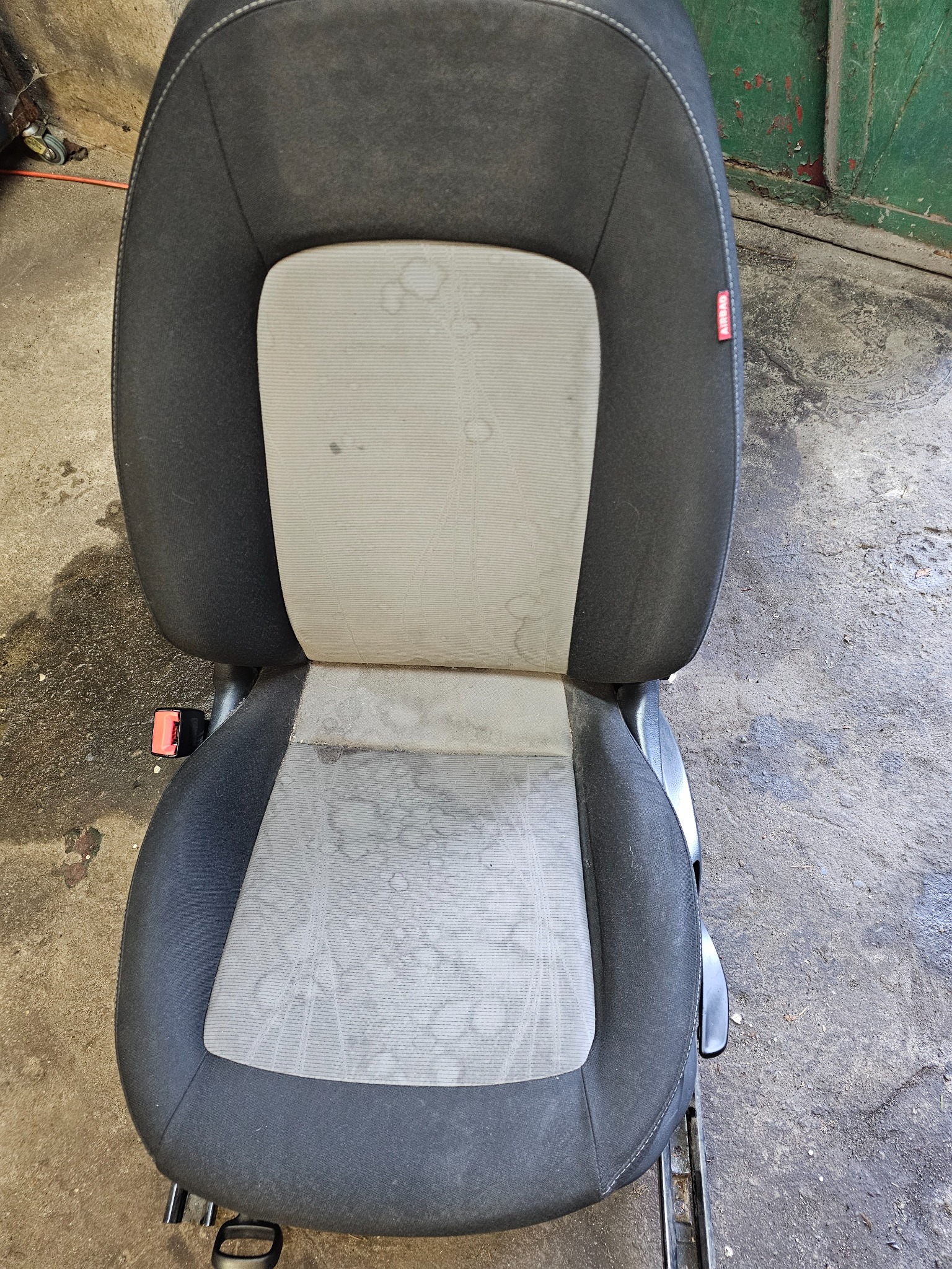 Widok z góry na pojedyncze, wyjęte z samochodu, szaro-czarne, mocno zabrudzone i poplamione siedzenie samochodowe z widoczną etykietą 'AIRBAG' i elementami mocowania na szynach.