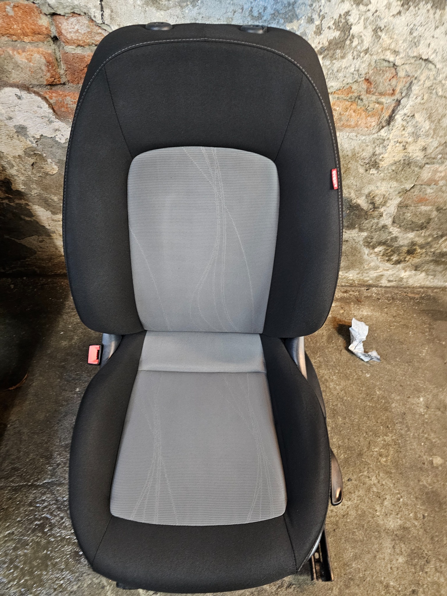 Fotel samochodowy z szarą tapicerką i czarnymi bokami, widoczny na tle ceglanej ściany i betonowej podłogi z leżącą chusteczką.