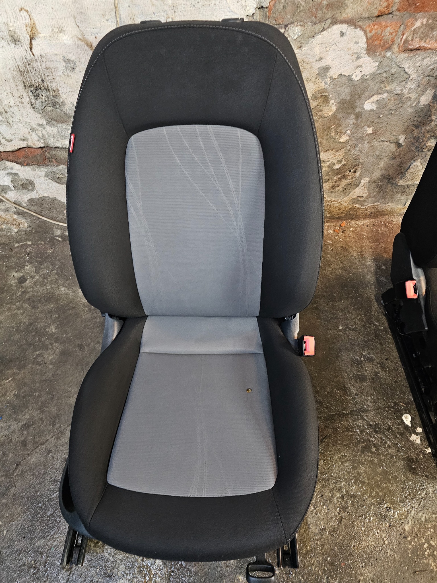 Wyjęte samochodowe siedzenie z szarą tapicerką i czarnym obramowaniem, widoczne ślady użytkowania, na tle surowej ściany i podłogi.