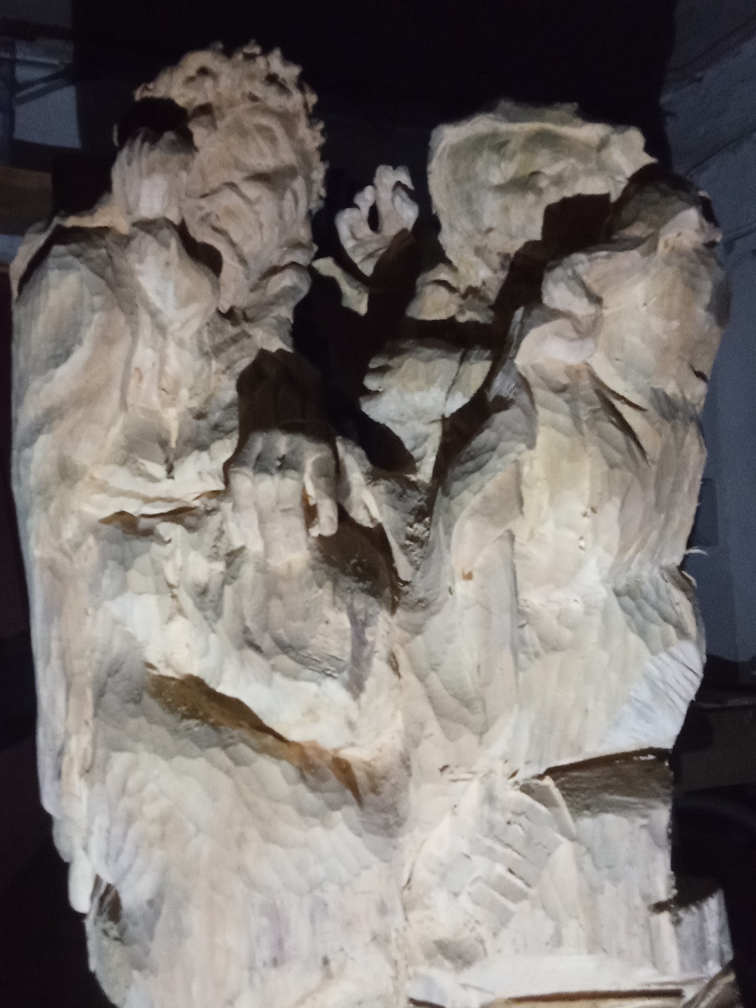 Rzeźba w drewnie przedstawiająca postacie, prawdopodobnie aniołów lub świętych, wykonana w Łowiczu, widoczna w słabym oświetleniu, z wyraźnymi śladami dłuta.