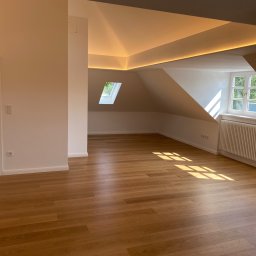 Bau-Remo usługi remontowo budowlane - Jasne, przestronne poddasze z drewnianą podłogą, oknami dachowymi i listwami LED wzdłuż krawędzi sufitu, świeżo po remoncie.
