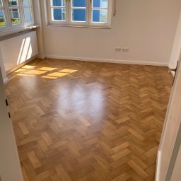 Bau-Remo usługi remontowo budowlane - Pokój po remoncie z parkietem ułożonym w jodełkę, białe ściany, okna z widokiem na niebieski budynek, promienie słoneczne na podłodze.