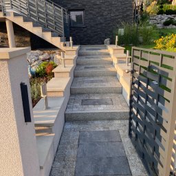 Bau-Remo usługi remontowo budowlane - Schody wejściowe do domu z szarej kostki brukowej, balustrady ze stali nierdzewnej, nowoczesne oświetlenie zewnętrzne i brama z metalowymi przęsłami.