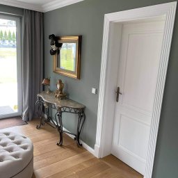 Bau-Remo usługi remontowo budowlane - Eleganckie wnętrze: oliwkowa ściana z dekoracyjnym lustrem, biała stolarka drzwiowa z frezowanymi listwami, konsola z marmurowym blatem i toczonymi nogami, pufa na drewnianej podłodze.