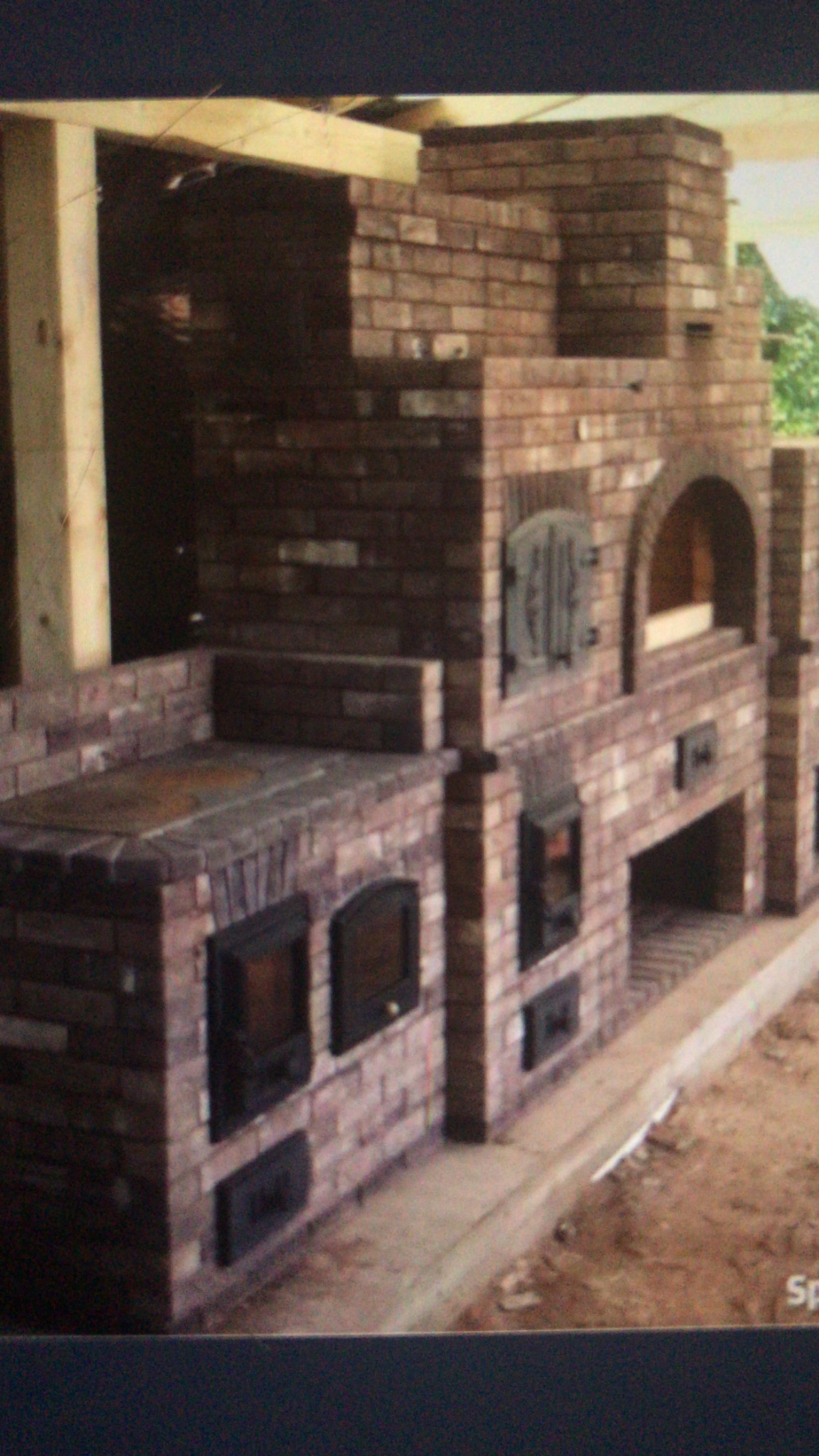 Murowana konstrukcja z cegły, przypominająca piec chlebowy lub wędzarnię, z drewnianą konstrukcją podtrzymującą, jeszcze w trakcie budowy, z widocznymi drzwiczkami.