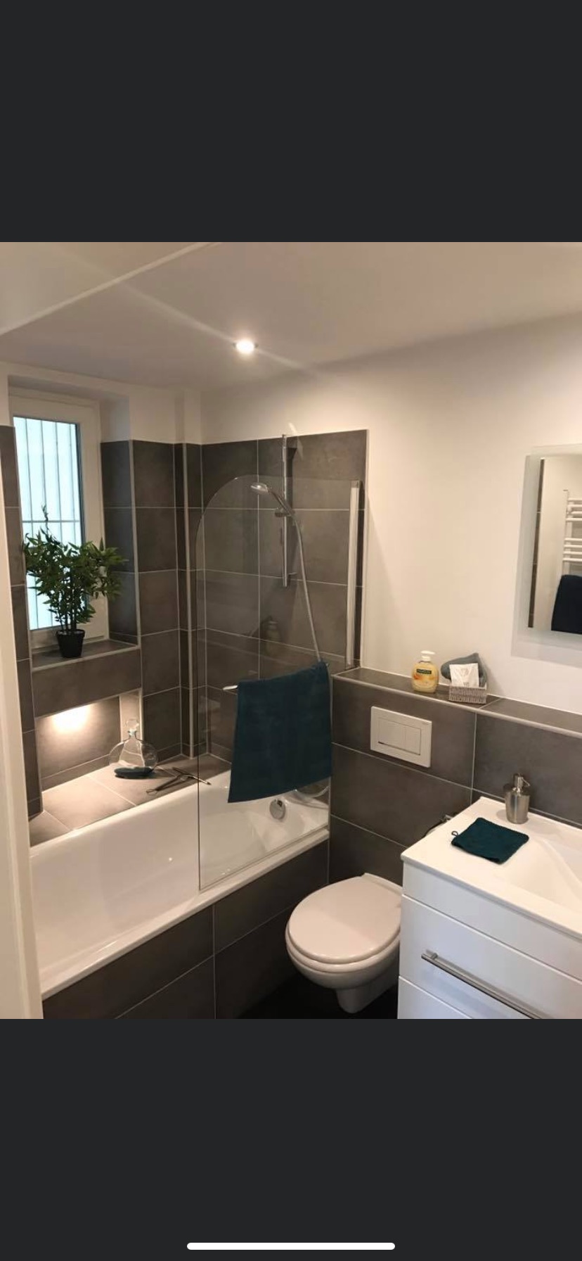 Nowoczesna łazienka z szarymi płytkami, białą armaturą, kabiną prysznicową z elementami szkła i rośliną w doniczce w niszy nad wanną.