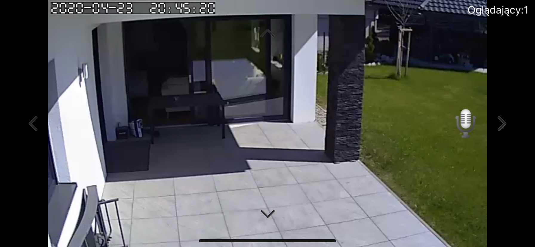 Nowoczesny taras z szarych płyt, widok z kamery monitoringu, z widocznym fragmentem trawnika i przeszklonym wejściem do domu.