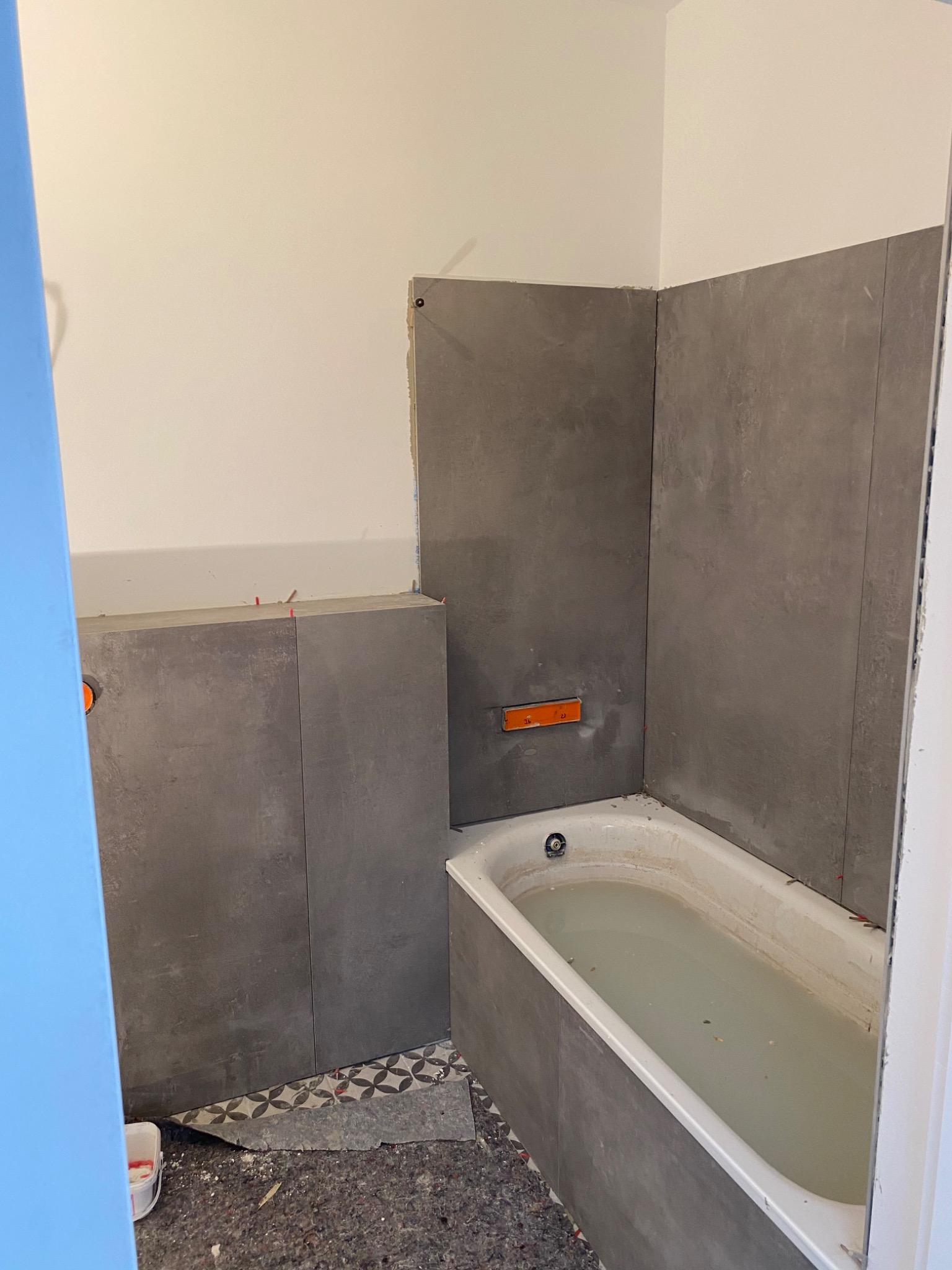 Łazienka w trakcie remontu: szare płytki imitujące beton częściowo ułożone wokół wanny wypełnionej brudną wodą, widoczne poziomice i krzyżyki dystansowe, podłoga zabezpieczona folią.