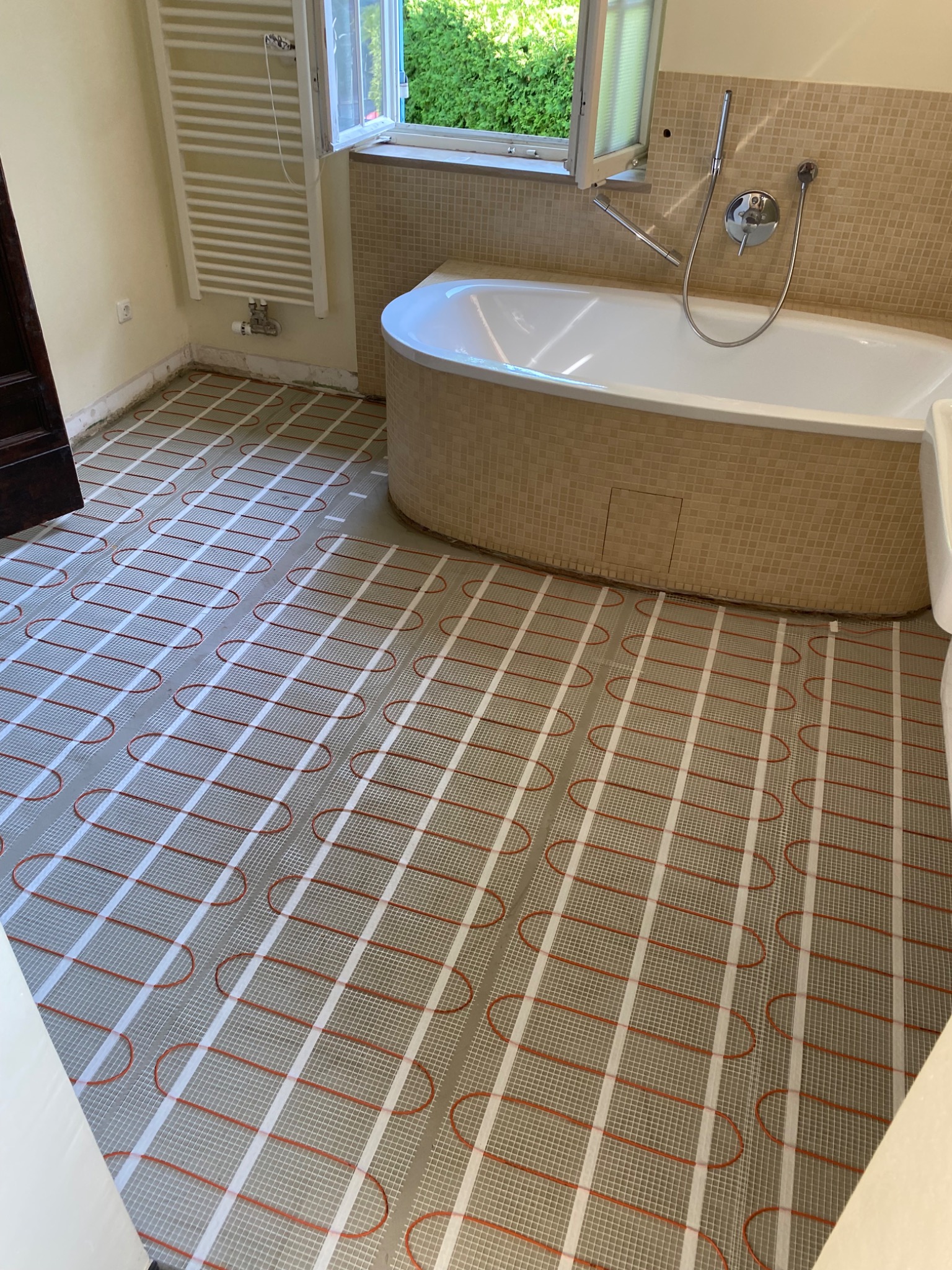 Biała wanna z mozaiką w łazience z zainstalowanym systemem ogrzewania podłogowego z czerwonymi kablami grzewczymi na siatce.