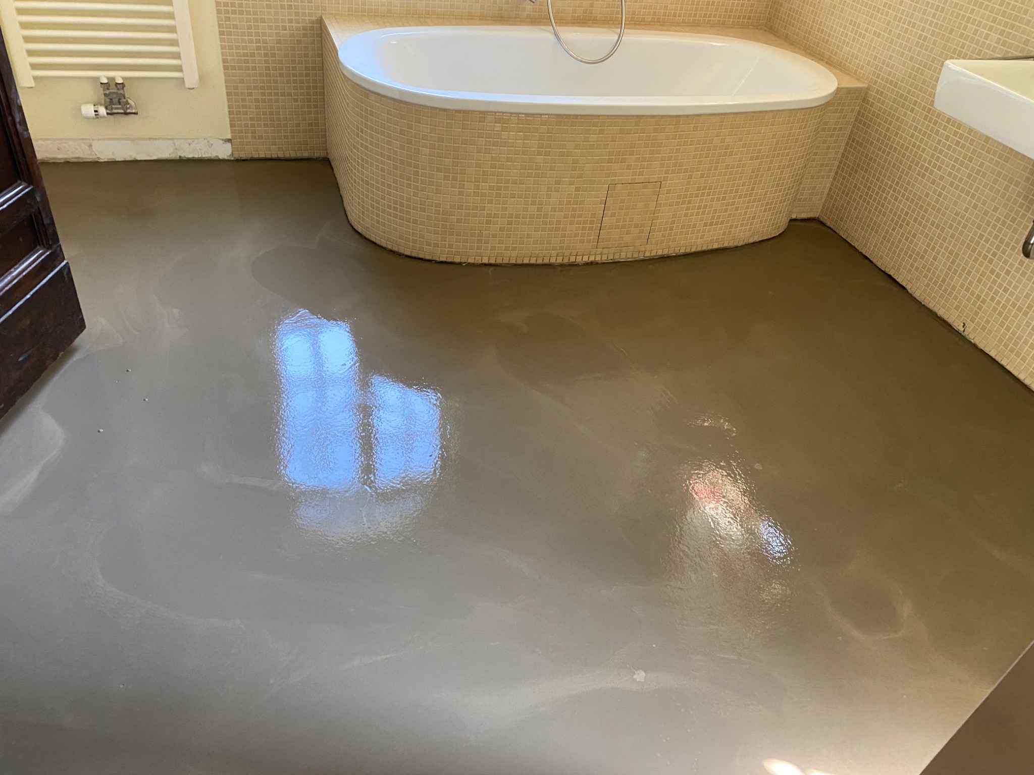 Świeżo wylana, beżowa posadzka żywiczna w łazience z wanną obudowaną mozaiką i częściowo wykończonymi ścianami. W posadzce odbija się okno.