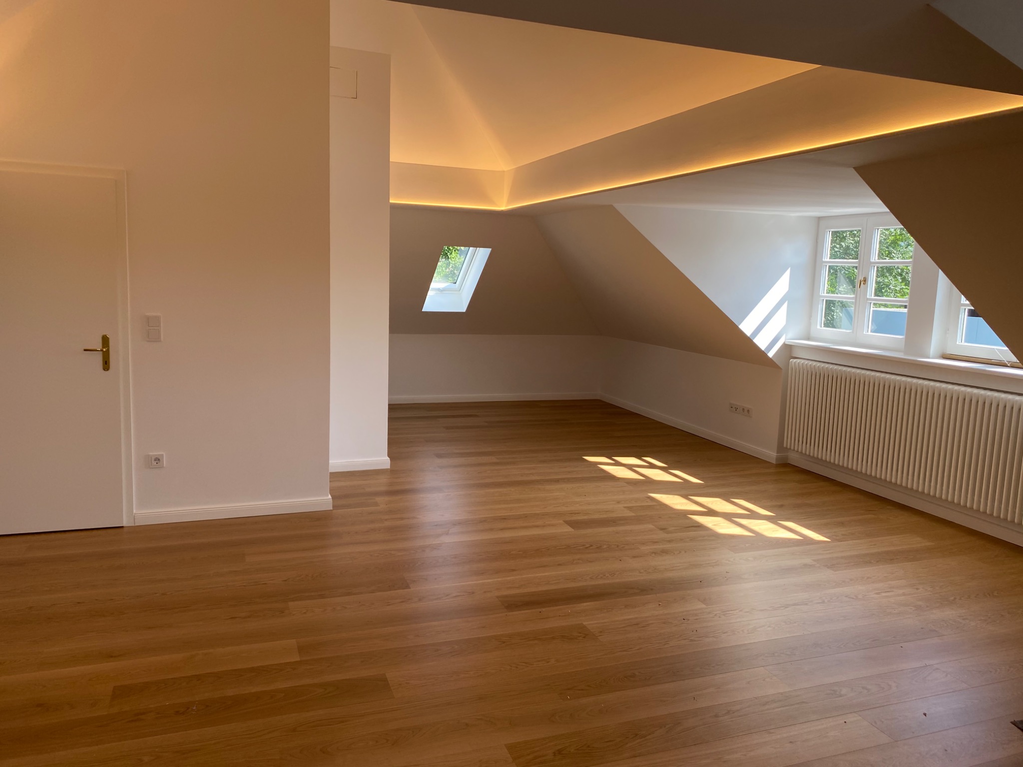 Jasne, przestronne poddasze z drewnianą podłogą, oknami dachowymi i listwami LED wzdłuż krawędzi sufitu, świeżo po remoncie.