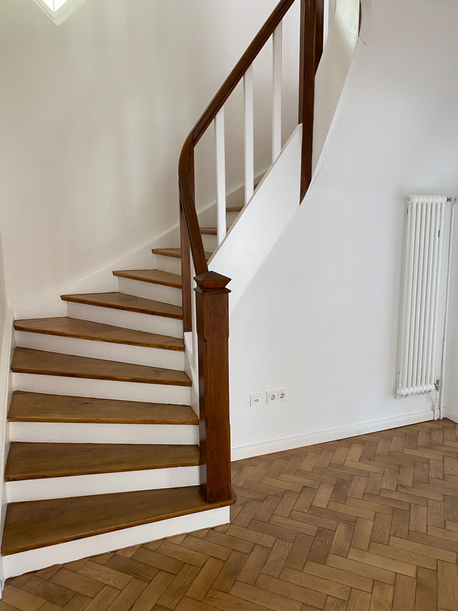 Drewniane schody z białymi podstopnicami i ciemnobrązową balustradą w jasnym wnętrzu, parkiet w jodełkę na podłodze, biały grzejnik na ścianie.
