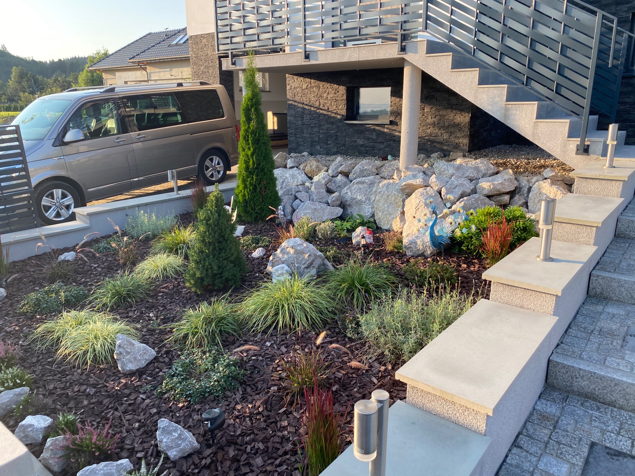 Ogród przydomowy z roślinami ozdobnymi, kamieniami, korą drzewną i dekoracyjną figurką pawia, widok z poziomu schodów wejściowych.