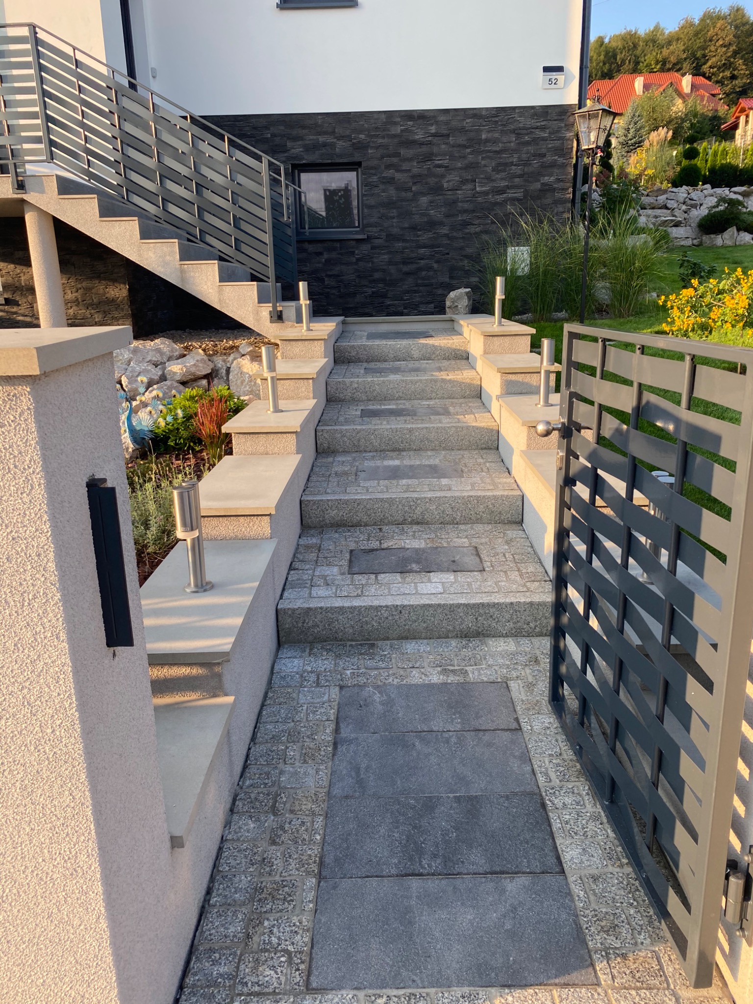 Schody wejściowe do domu z szarej kostki brukowej, balustrady ze stali nierdzewnej, nowoczesne oświetlenie zewnętrzne i brama z metalowymi przęsłami.