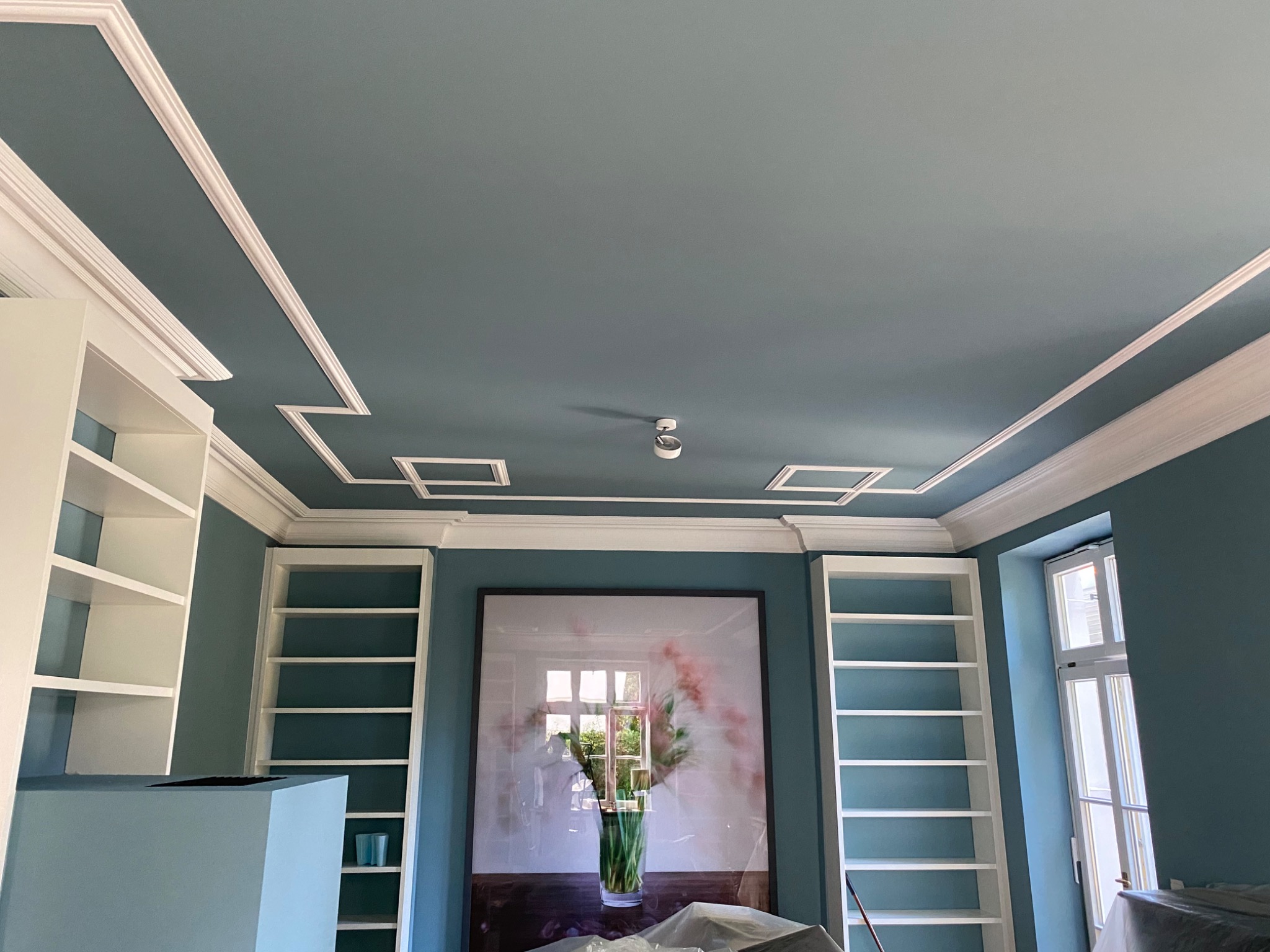 Turkusowy sufit z ozdobnymi białymi listwami i wbudowanym oświetleniem, ściany w tym samym odcieniu turkusu, białe regały wnękowe po bokach i obraz w ramie na środku