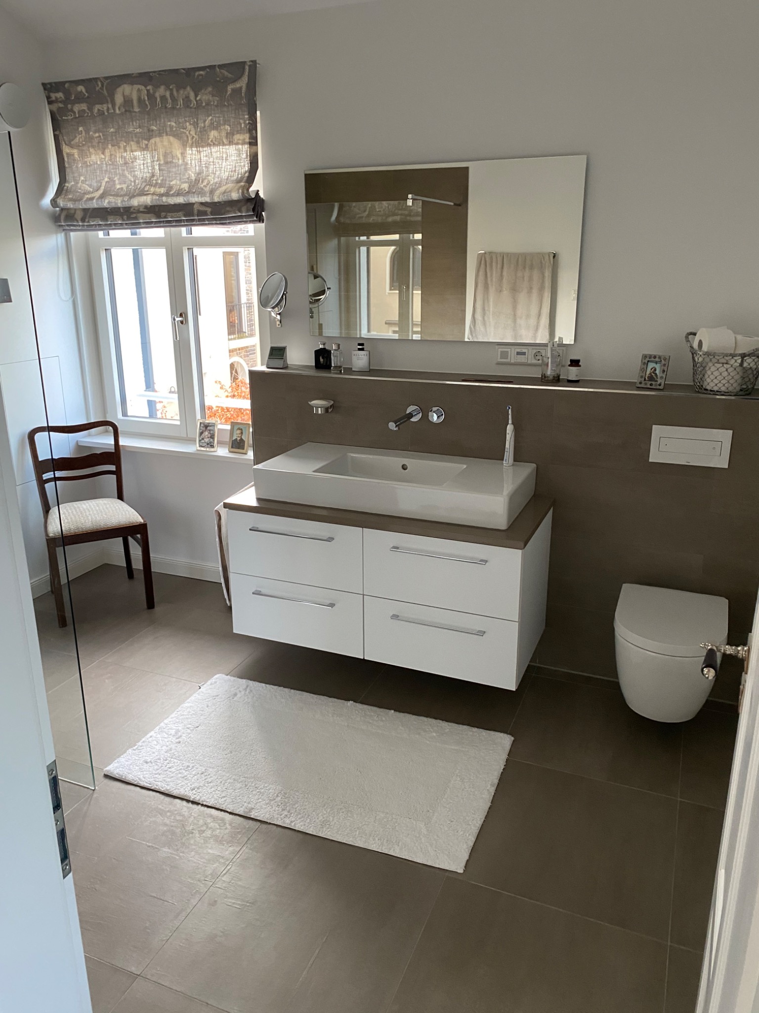 Nowoczesna łazienka z białą szafką pod umywalką, lustrem, toaletą i oknem z zasłoną w motywy zwierzęce, brązowe płytki podłogowe.