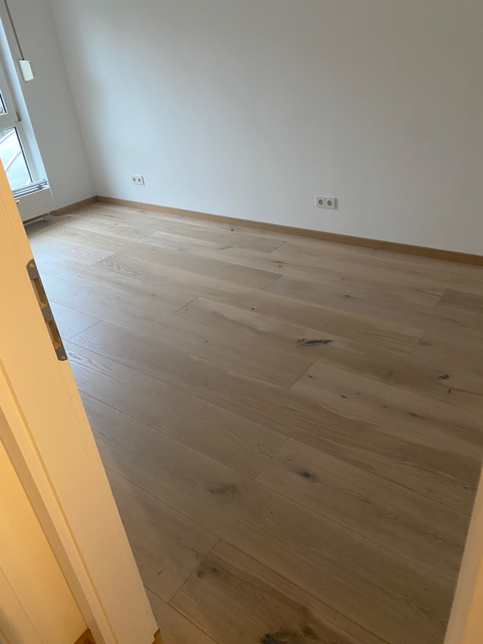 Pusty pokój z parkietem o naturalnym odcieniu drewna, świeżo pomalowane białe ściany, widoczne gniazdka elektryczne i fragment otwartych drzwi po lewej stronie.