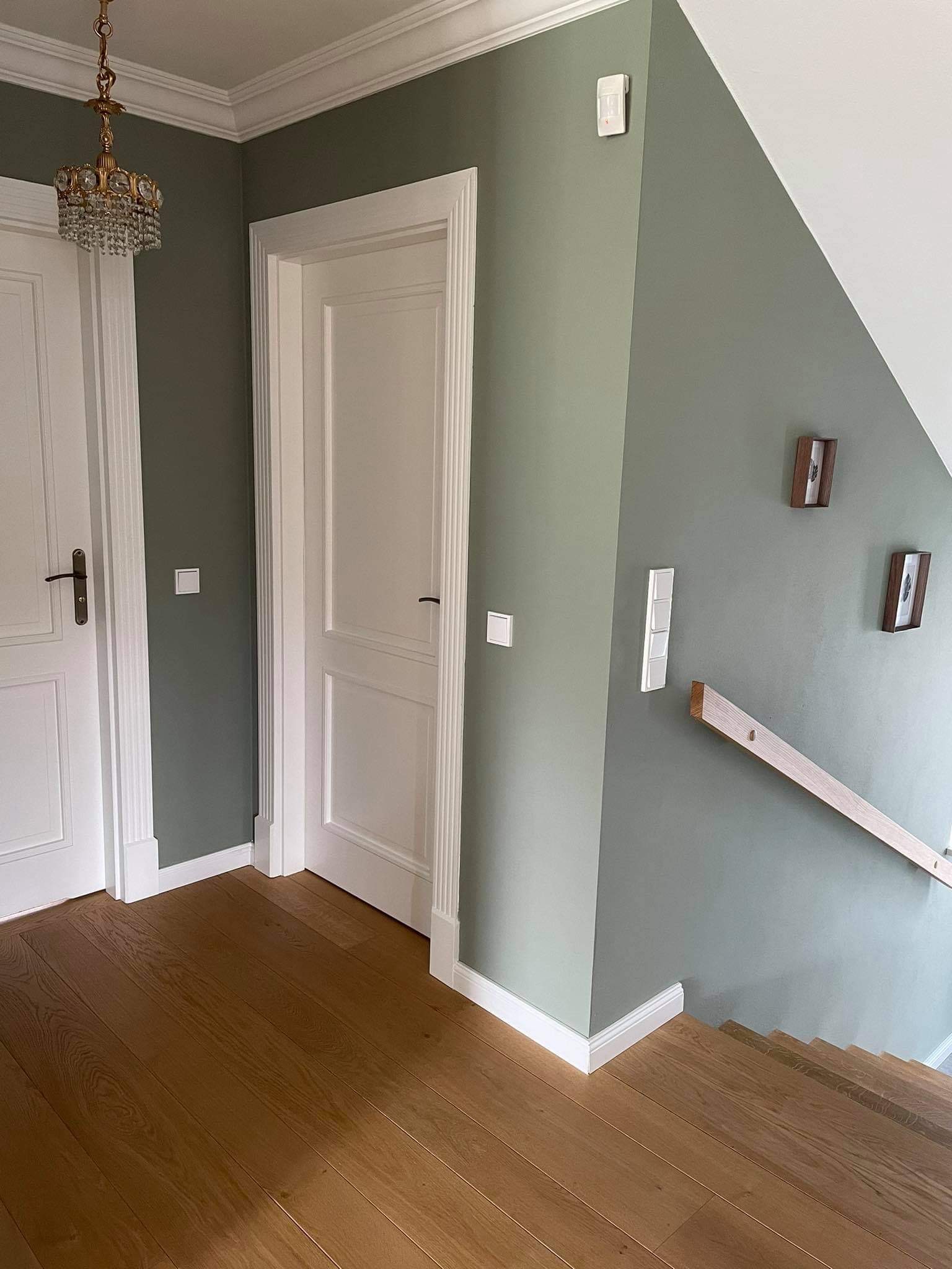 Korytarz w domu z drewnianą podłogą, ścianami w kolorze oliwkowej zieleni, białymi drzwiami z ozdobnymi framugami i schodami z drewnianą balustradą. Na ścianie wisi kryształowy żyrandol i drewniane...